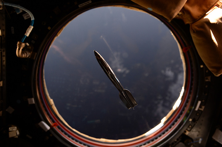 The Mini Hugo Rocket floats in the International Space Station in front of a window overlooking the Earth.