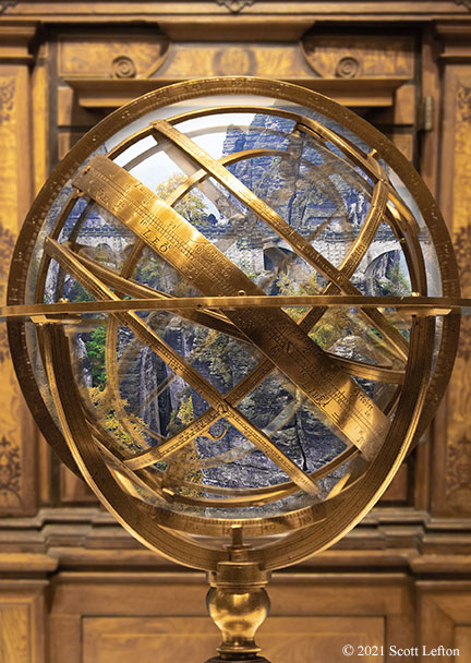 An armillary sphere in front of an ornate cabinet, where within the sphere is a vision of a far-away landscape with fortifications. 
