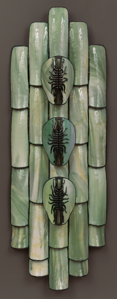 A stained glass sculpture, mostly in green with some white and yellow. The glass pieces resemble long, thin ceramic roof tiles. In front of the main sculpture are three stained glass masks with central bronze castings having insectlike tendril motifs.
